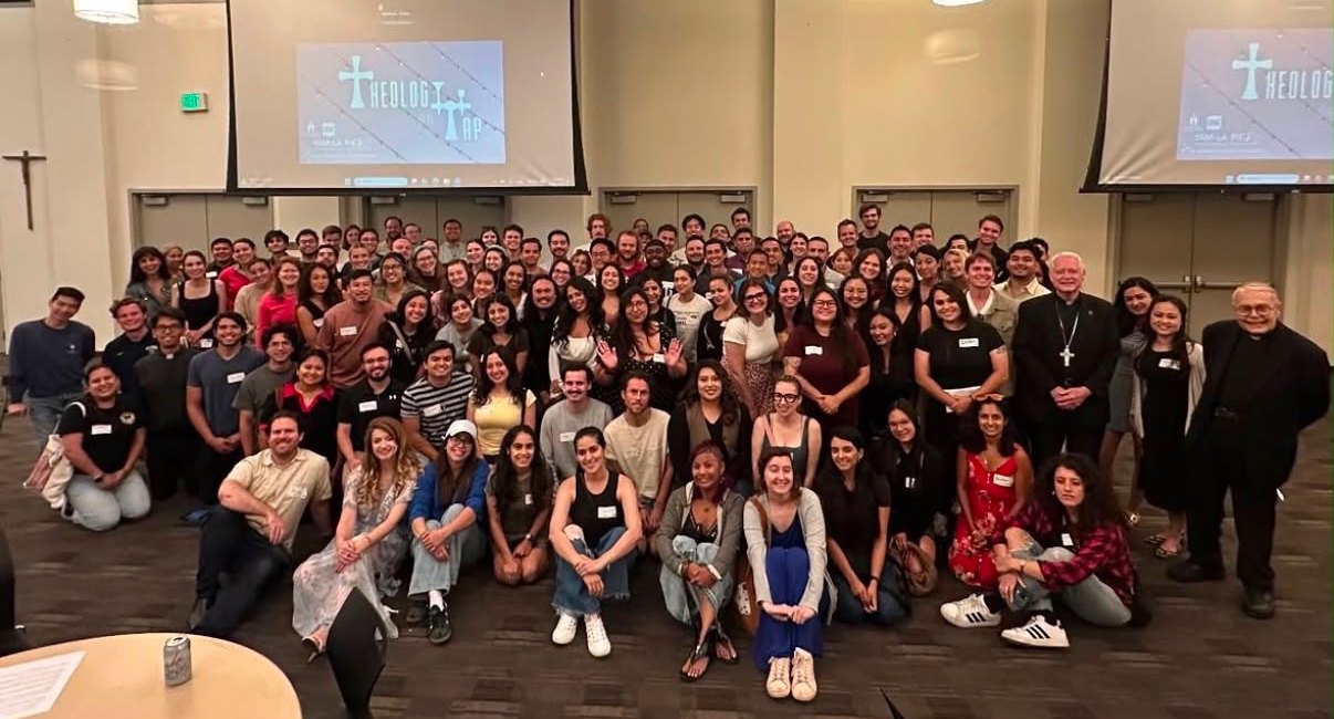 A full room of young adults at Theology on Tap, sitting on the floor and standing in rows, two priests among them, the Theology on Tap slide on the screens behind